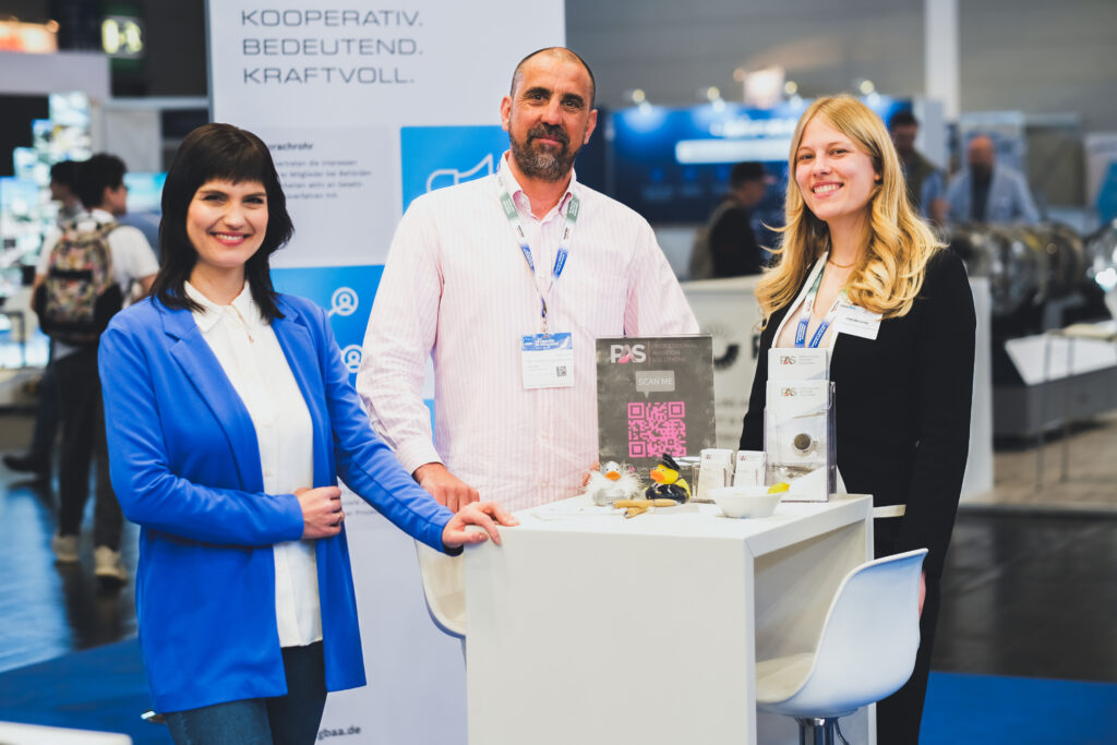 Drei Personen auf der AERO 2025 in Friedrichshafen, versammelt um einen Tisch, lächeln in die Kamera.